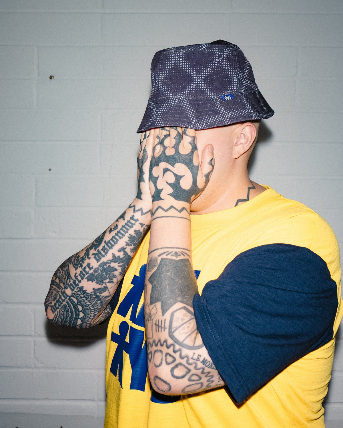Lockwood Avenue ADWYSD Human T-Shirt in Yellow/Navy worn by a tattooed model with a bucket hat pulled down, hands covering the face