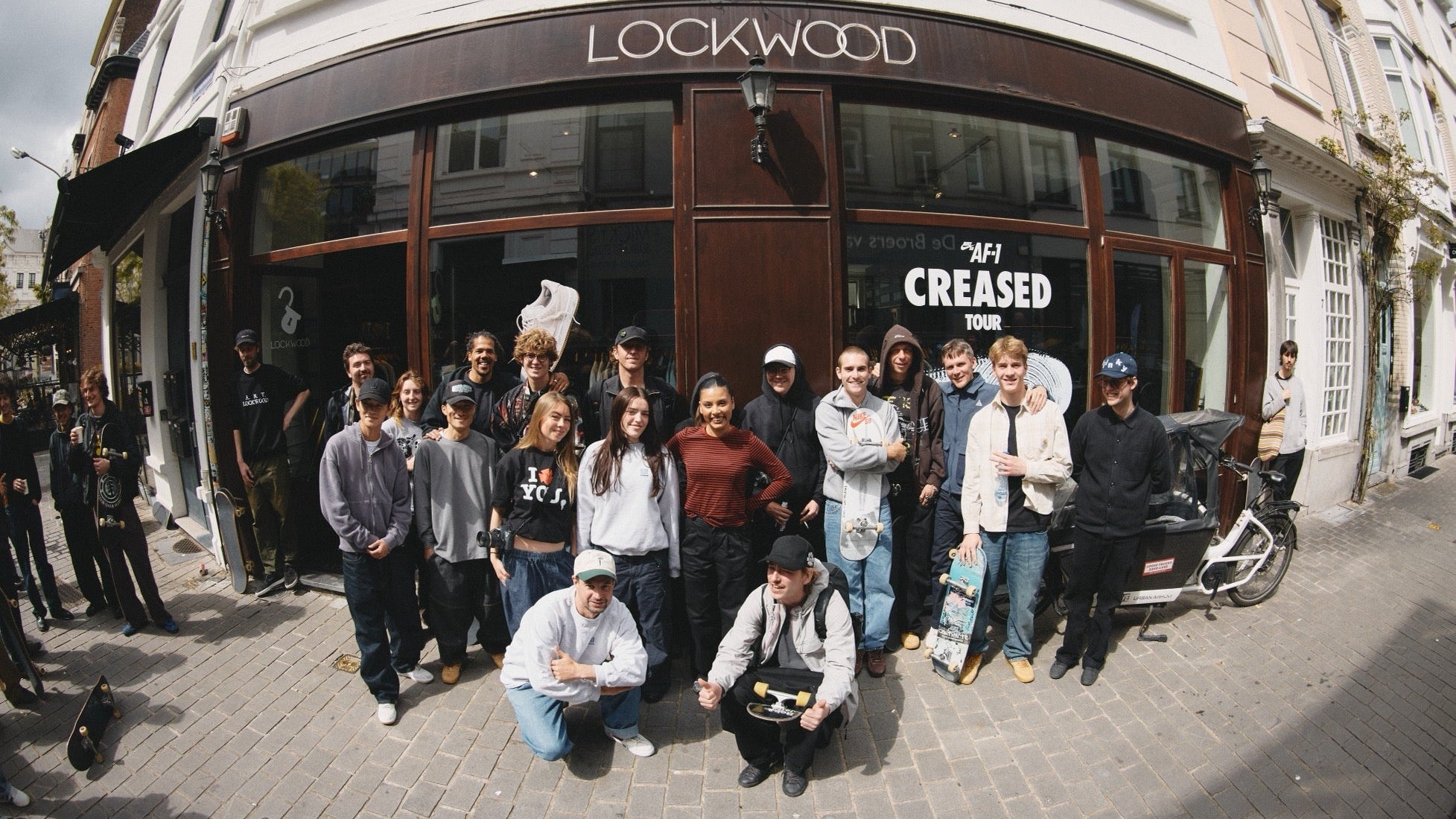 A group of people posing in front of a Lockwood storefront on a city street. The shop has large windows, a dark brown awning, and a white facade.