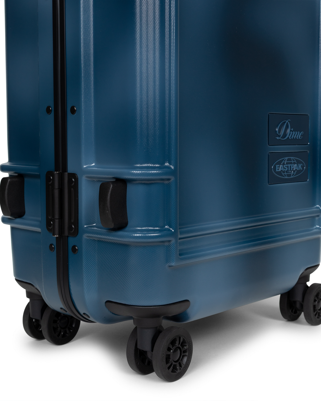 Close up of the lower corner of the Eastpak x Dime Resist'R Case S in Dime Navy, showing two of the four spinner wheels, the moulded corner bumpers and the Dime and Eastpak badges