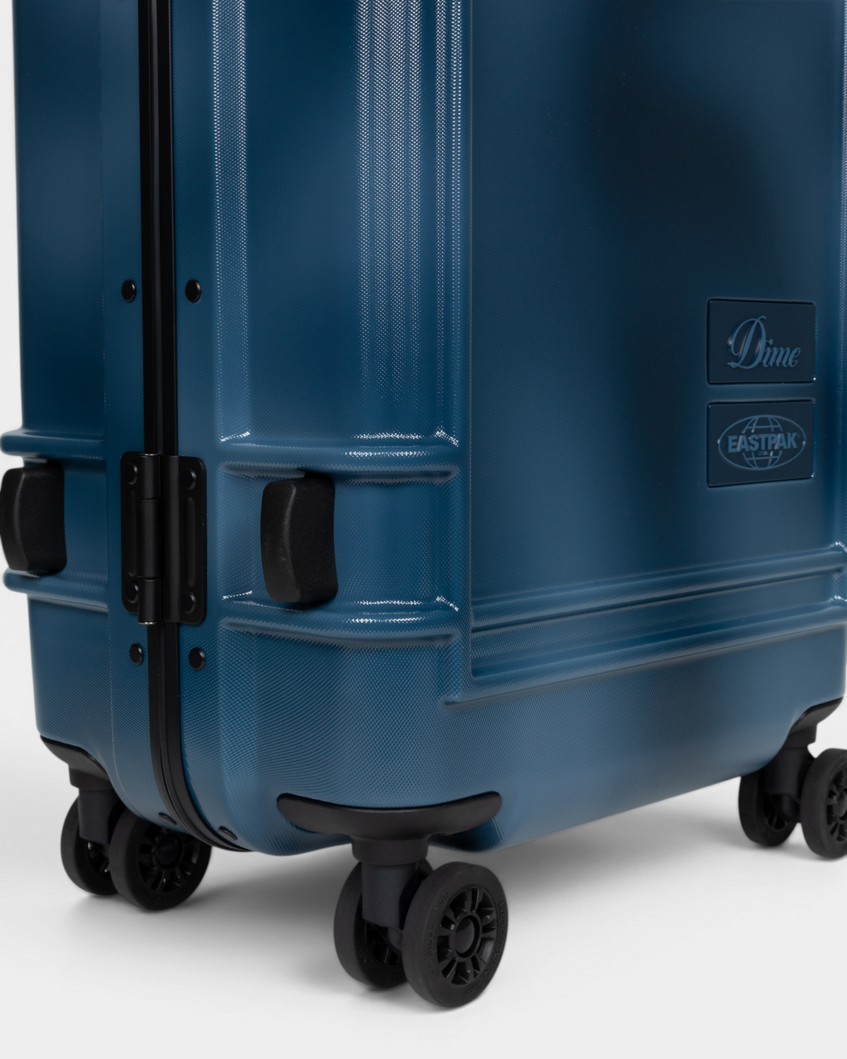 Close up of the lower corner of the Eastpak x Dime Resist'R Case S in Dime Navy, showing two of the four spinner wheels, the moulded corner bumpers and the Dime and Eastpak badges