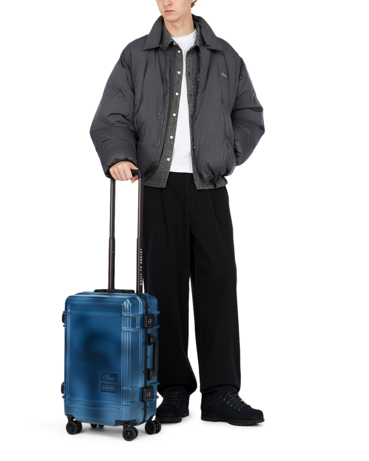 A person in a grey puffer jacket wheeling the Eastpak x Dime Resist'R Case S in Dime Navy by its extended telescopic handle, showing the cabin size against a standing adult