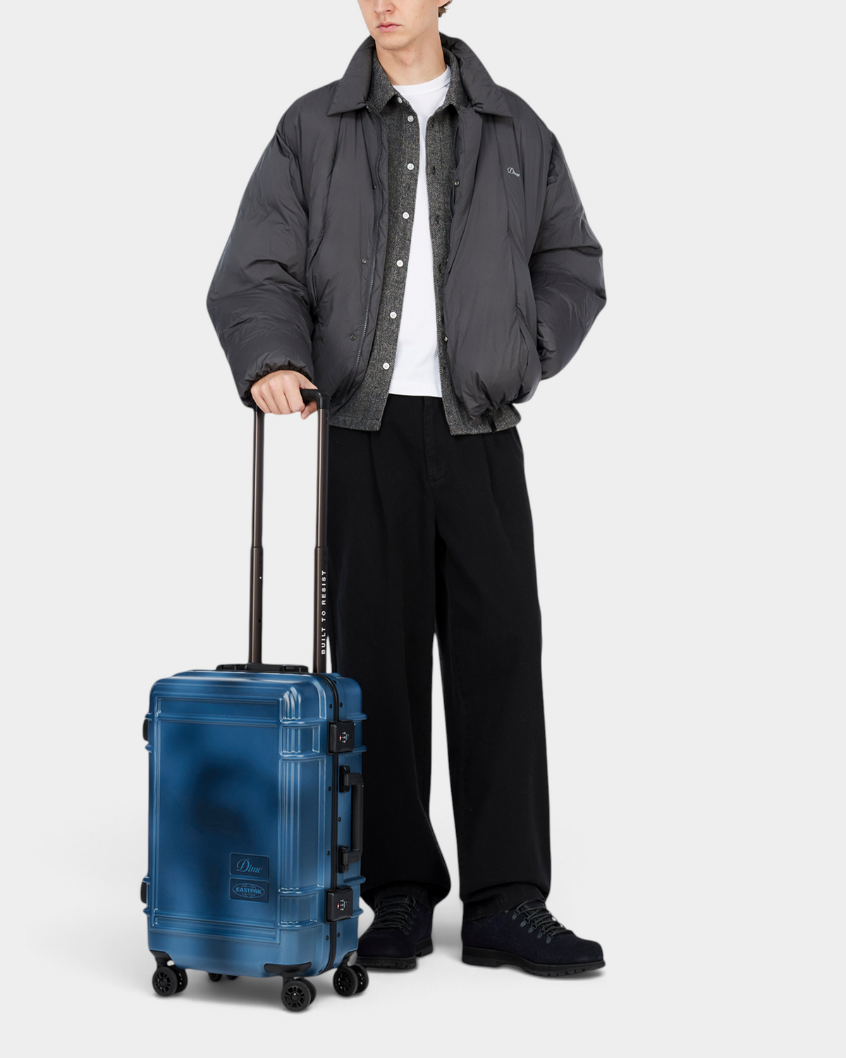 A person in a grey puffer jacket wheeling the Eastpak x Dime Resist'R Case S in Dime Navy by its extended telescopic handle, showing the cabin size against a standing adult