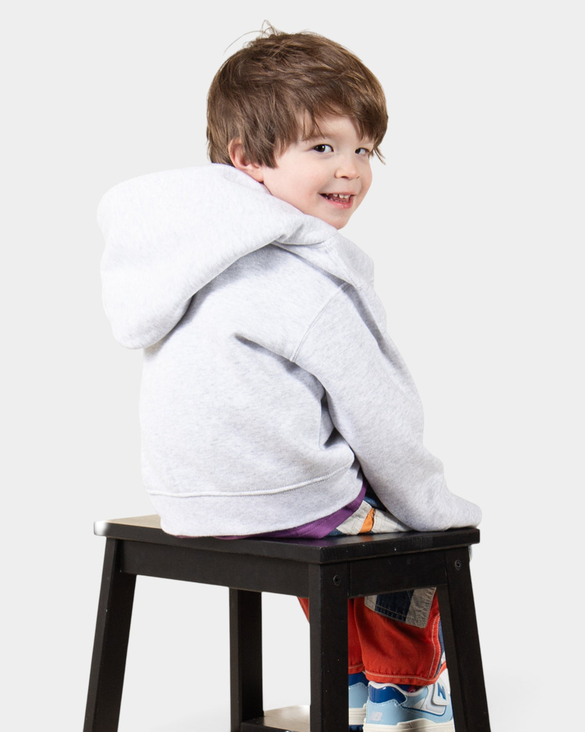A young boy wearing a Lockwood Avenue ash gray Dime fleece hoodie while sitting on a black stool.