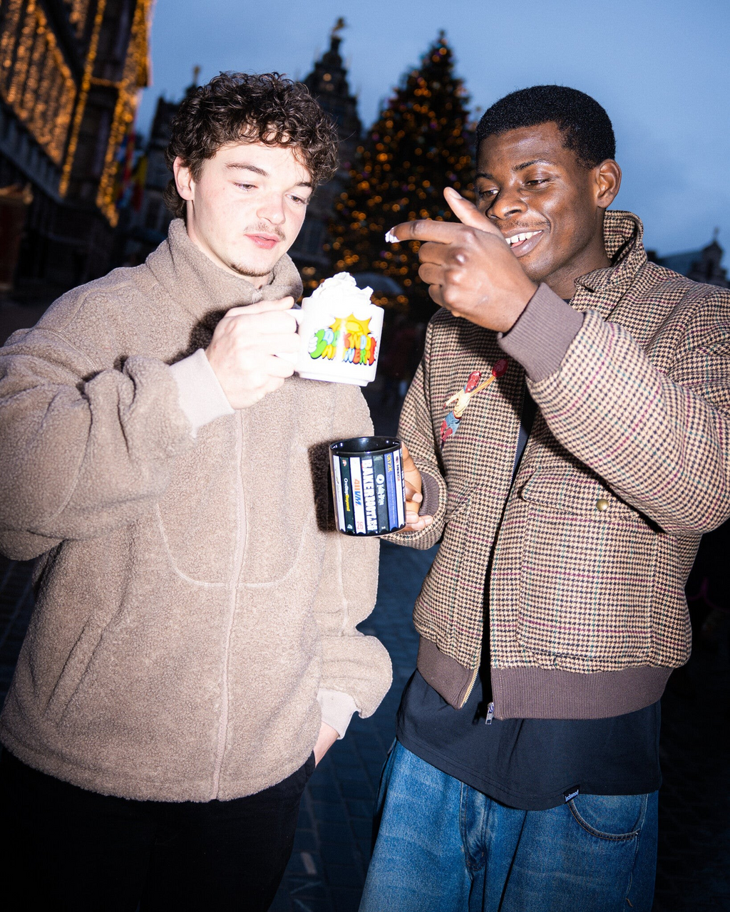 Two men holding Lockwood Avenue ceramic mugs with Sunny Antwerp graphic prints outdoors in a city square at night.
