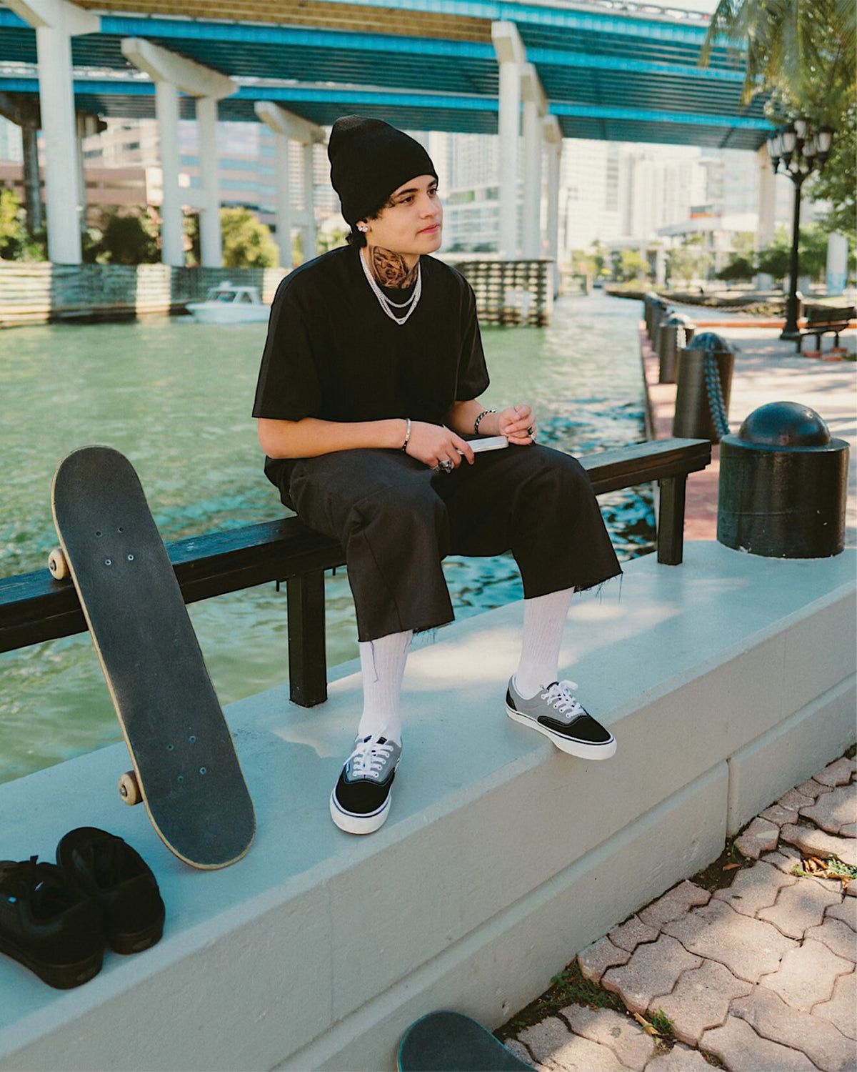 Lockwood Avenue model wearing black and grey Vans Skate Era low-top sneakers while sitting by a canal with a skateboard