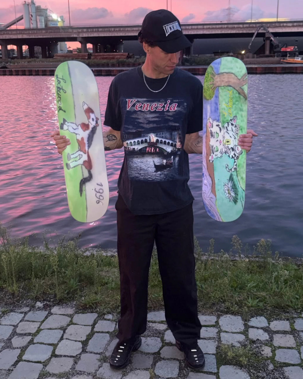 Artist Attila Buonsenso standing by the water at sunset holding both Lockwood x Attila decks, the tiger cub graphic in his left hand