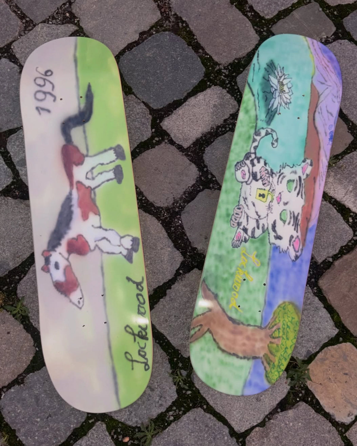 The two Lockwood x Attila decks lying side by side on Antwerp cobblestones, the tiger cub graphic on the right and the horse on the left