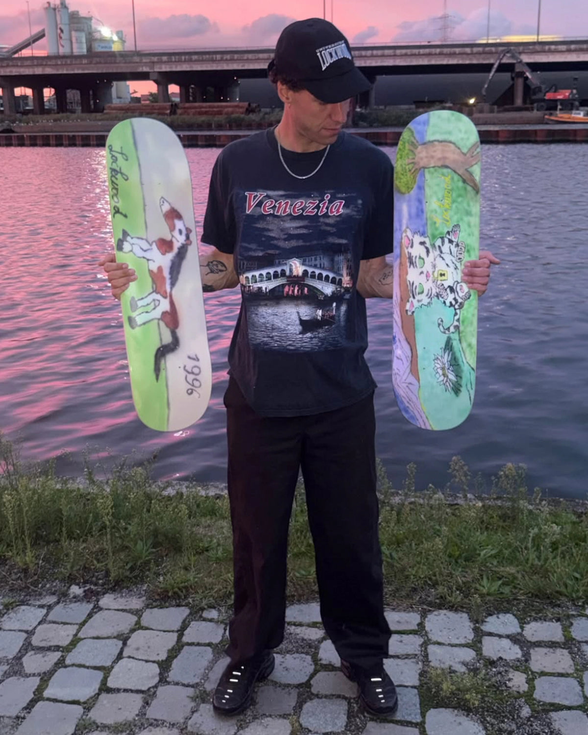 Artist Attila Buonsenso standing by the water at sunset holding both Lockwood x Attila decks, the tiger cub graphic in his left hand