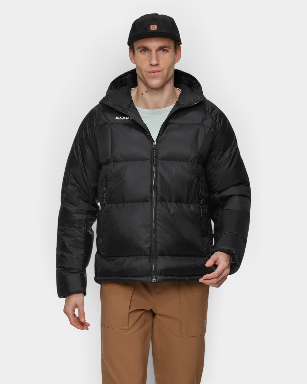 A man wearing the Mammut Glacier Glow IN Hooded Jacket in black open over a pale tee, hood down behind the collar, with tan Mammut trousers