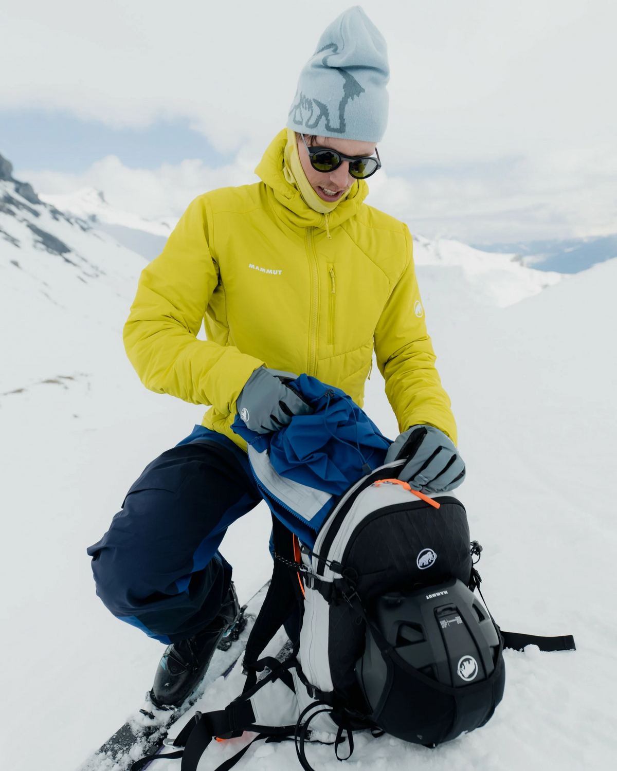 A skier on a snow slope in Mammut kit, wearing the pale Strata side of the reversible logo beanie with a yellow insulated jacket and a Mammut pack