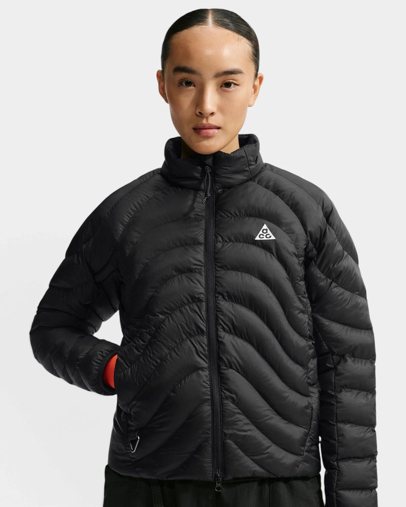 Nike ACG Lava Flow Women's Jacket in black and summit white, front view on a model with the stand collar zipped up, wavy quilted baffles across the body and the white ACG triangle logo on the chest