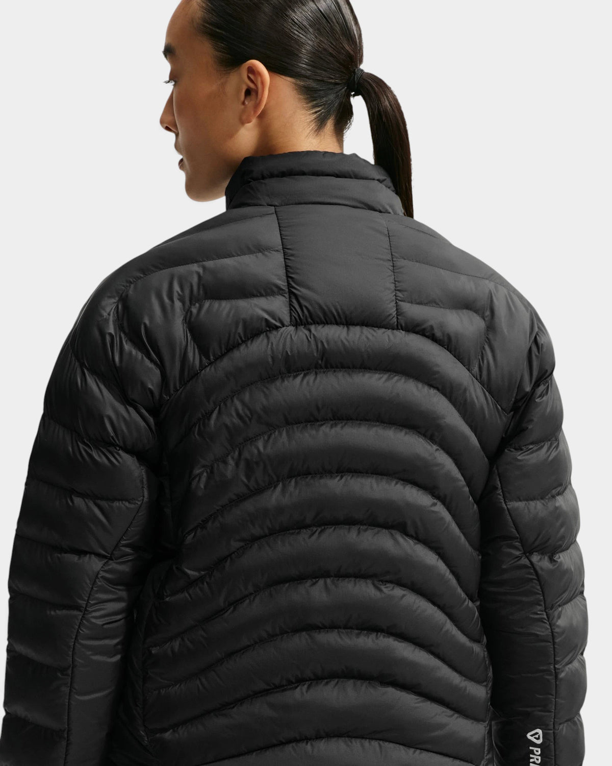 Nike ACG Lava Flow Women's Jacket in black and summit white, three-quarter back view on a model, showing the curved quilted baffles across the back and the PrimaLoft label on the lower seam