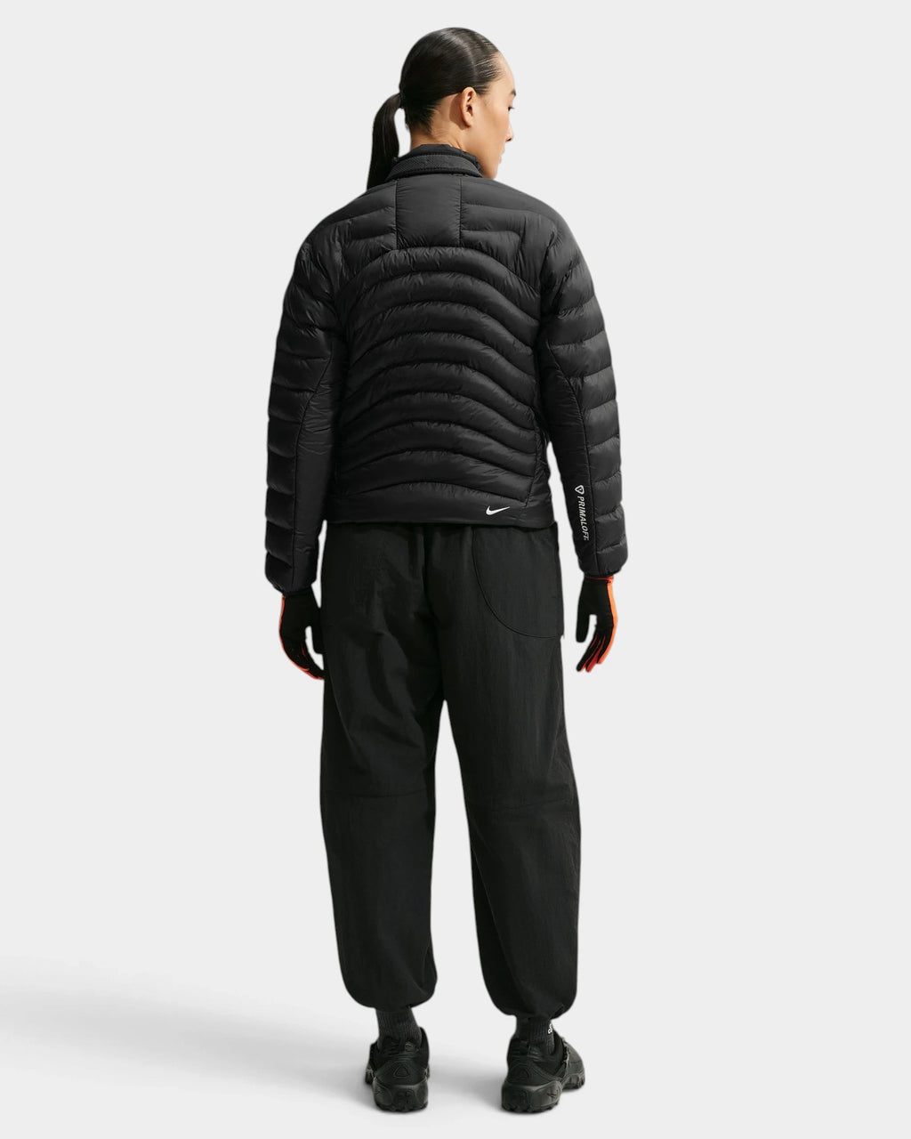 Nike ACG Lava Flow Women's Jacket in black and summit white, full back view on a model in black trousers, showing the quilted back panel and the small Nike swoosh at the hem