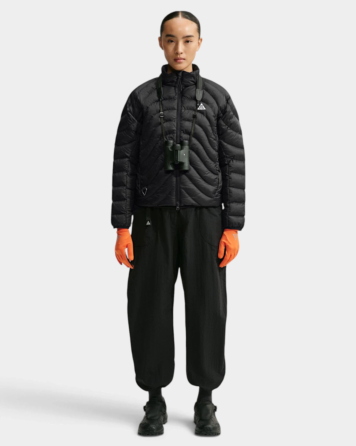 Nike ACG Lava Flow Women's Jacket in black and summit white, full-length front view on a model wearing black trousers, orange gloves and binoculars on a strap