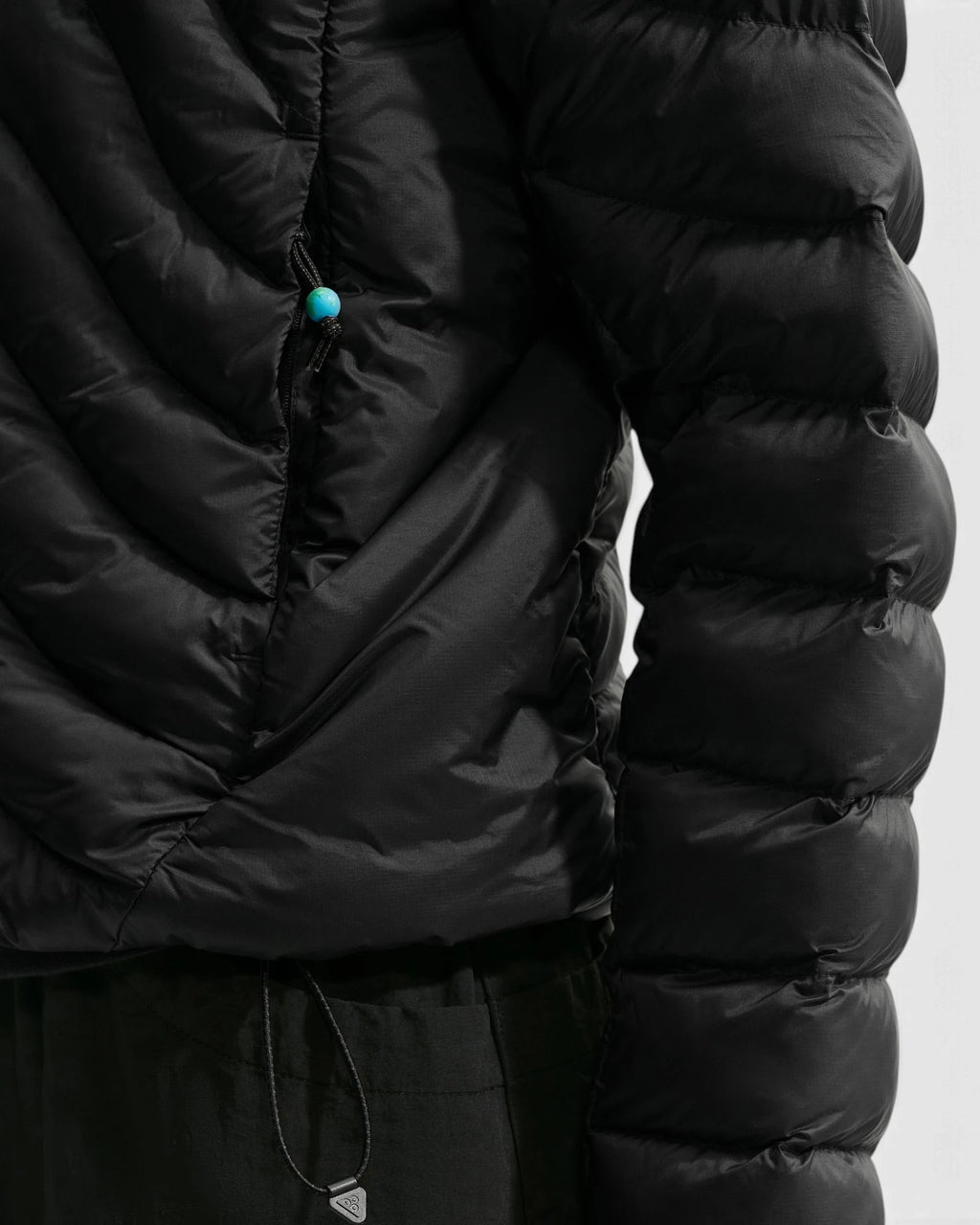 Close up of the Nike ACG Lava Flow Women's Jacket in black and summit white, showing the quilted sleeve, the zip pocket and the waist bungee cord with its turquoise toggle