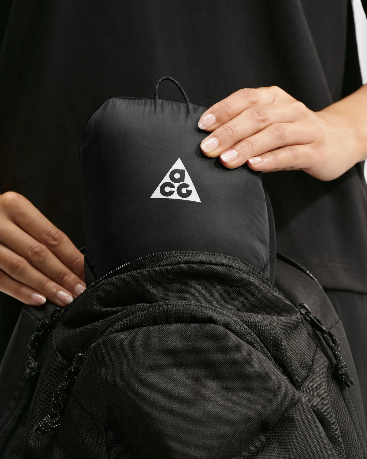 The Nike ACG Lava Flow Women's Jacket in black and summit white packed into its internal pocket and held against a black rucksack, with the ACG triangle logo on the pouch