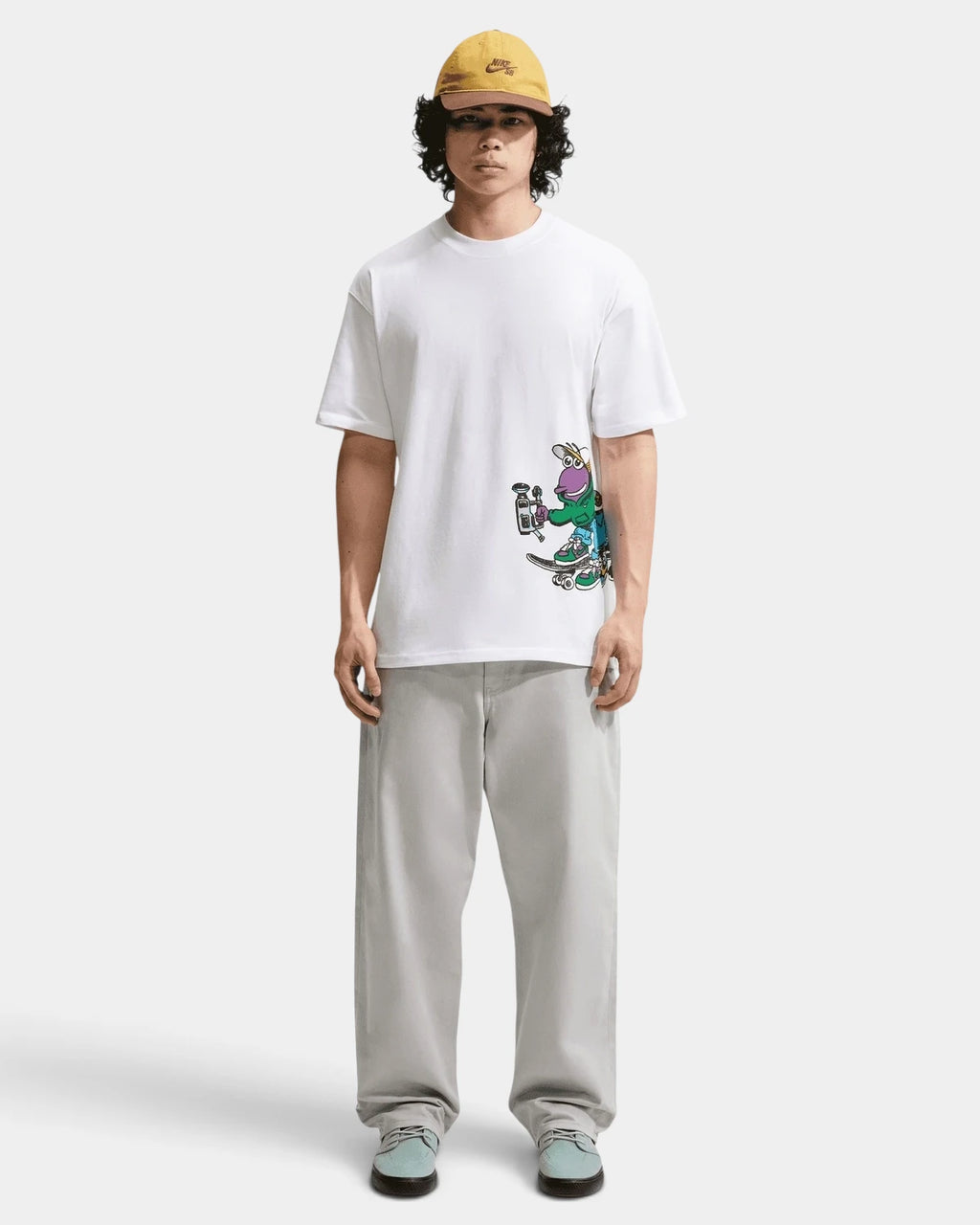 Full-length front view of a model in the white Nike SB Sk8pillar T-Shirt with grey trousers and a yellow cap