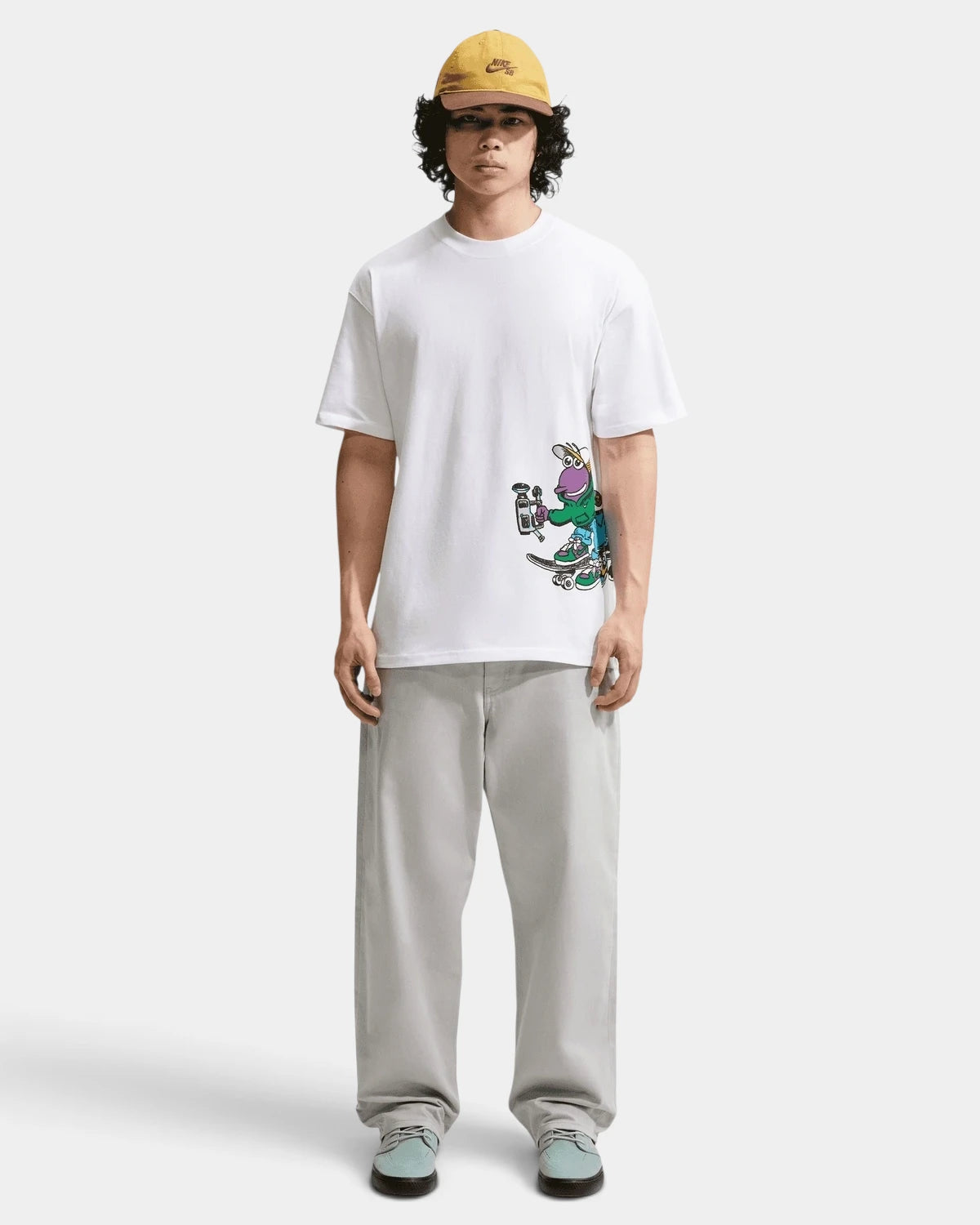 Full-length front view of a model in the white Nike SB Sk8pillar T-Shirt with grey trousers and a yellow cap