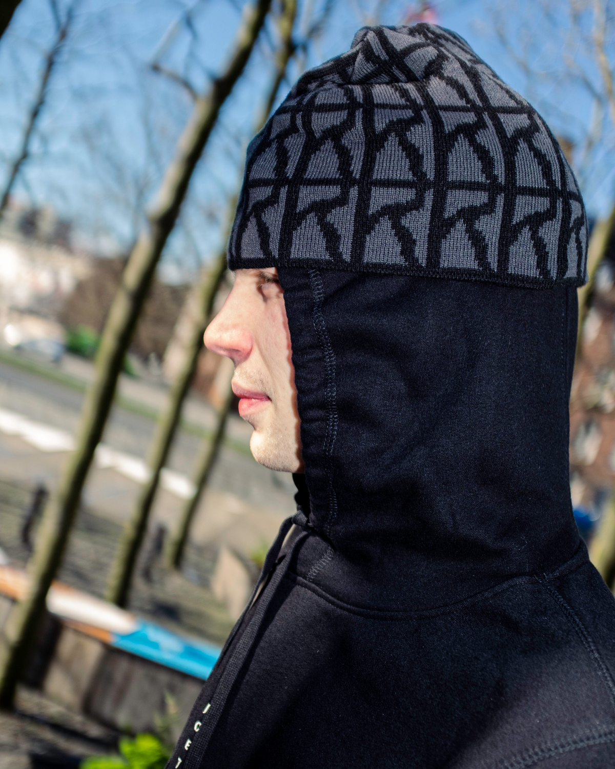 Lockwood Avenue Poetic Collective Skull Beanie in Logo Pattern worn by a model in profile, outdoors, over a black hood