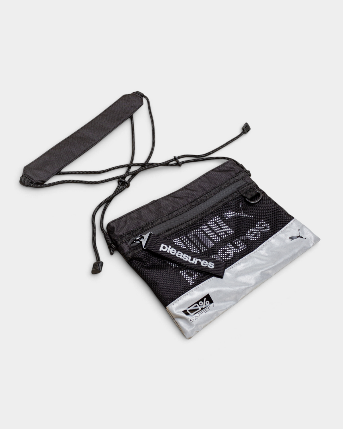 Puma x PLEASURES Body Bag in black, the whole flat crossbody laid out front on with its adjustable cord strap and removable padded shoulder pad beside it, showing the black mesh front panel, the reflective silver base and the Puma cat logo