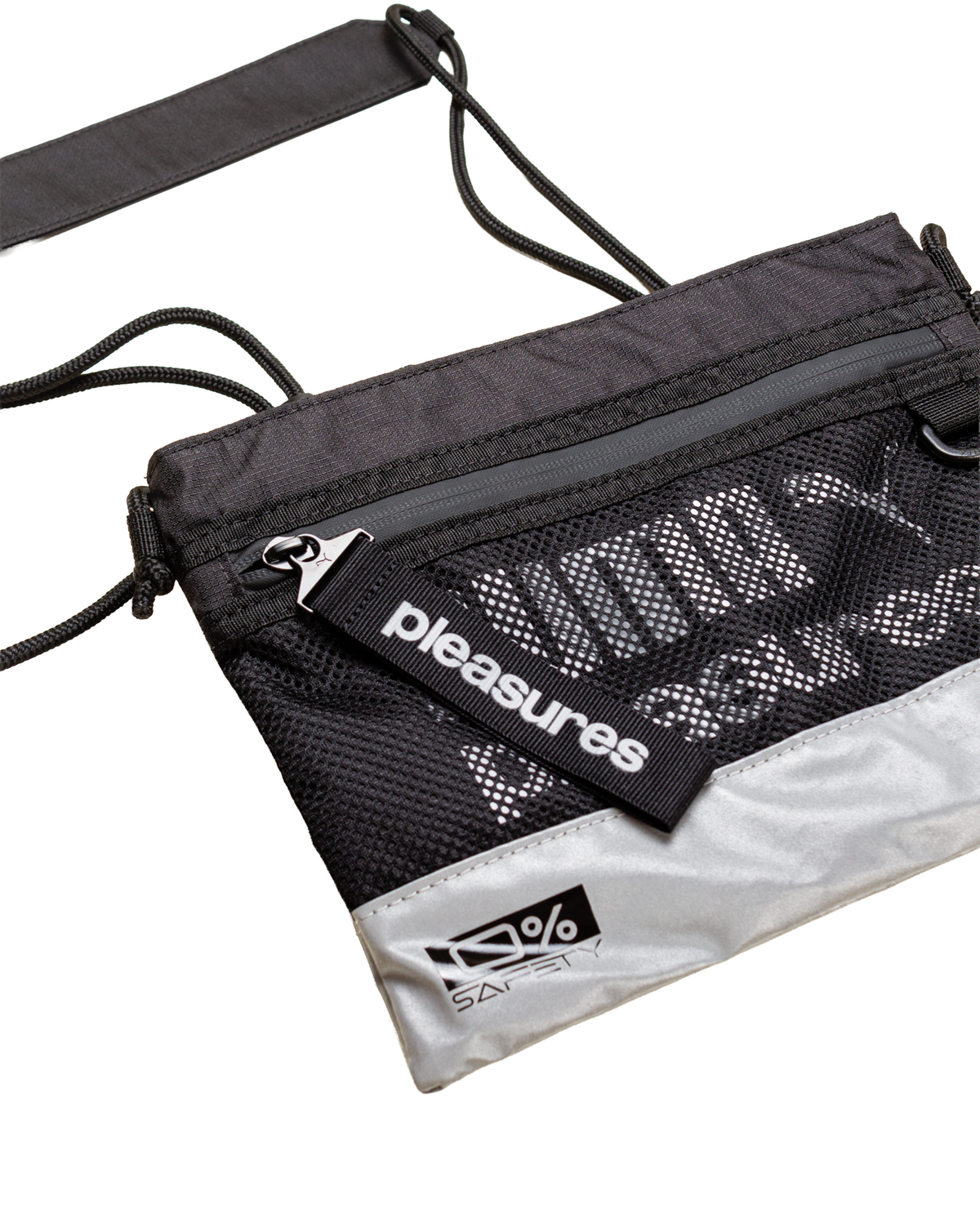 Puma x PLEASURES Body Bag in black seen from a three-quarter angle, showing the grey zip across the top, the open mesh over the printed panel, the PLEASURES webbing tab at the zip pull and the 0% SAFETY badge on the reflective silver base