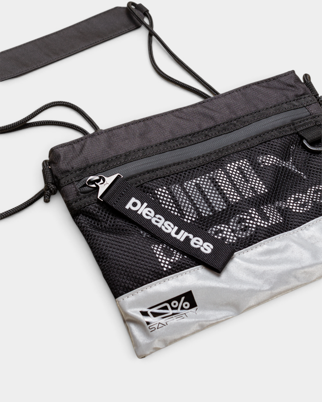 Puma x PLEASURES Body Bag in black seen from a three-quarter angle, showing the grey zip across the top, the open mesh over the printed panel, the PLEASURES webbing tab at the zip pull and the 0% SAFETY badge on the reflective silver base