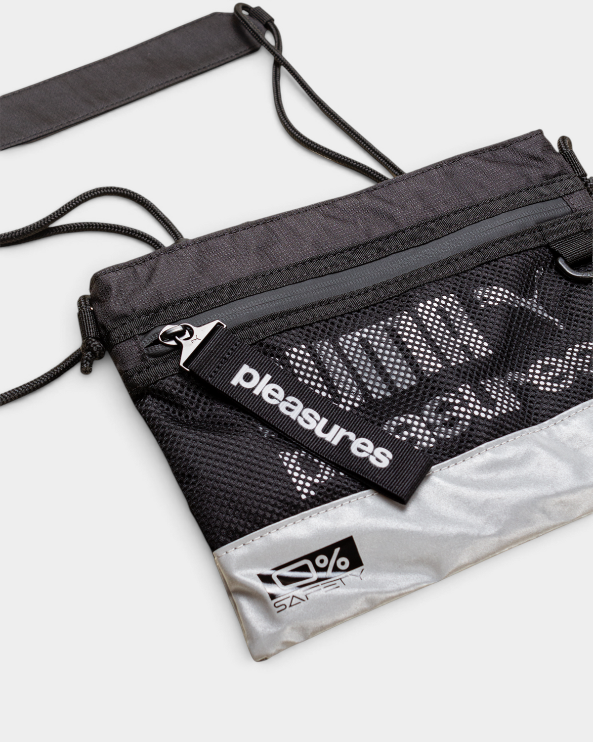 Puma x PLEASURES Body Bag in black seen from a three-quarter angle, showing the grey zip across the top, the open mesh over the printed panel, the PLEASURES webbing tab at the zip pull and the 0% SAFETY badge on the reflective silver base