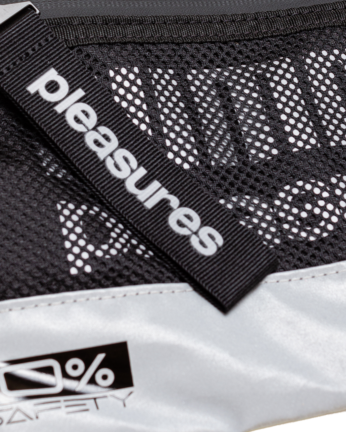Close up of the Puma x PLEASURES Body Bag in black, the PLEASURES webbing tab lying across the open mesh front panel above the reflective silver base
