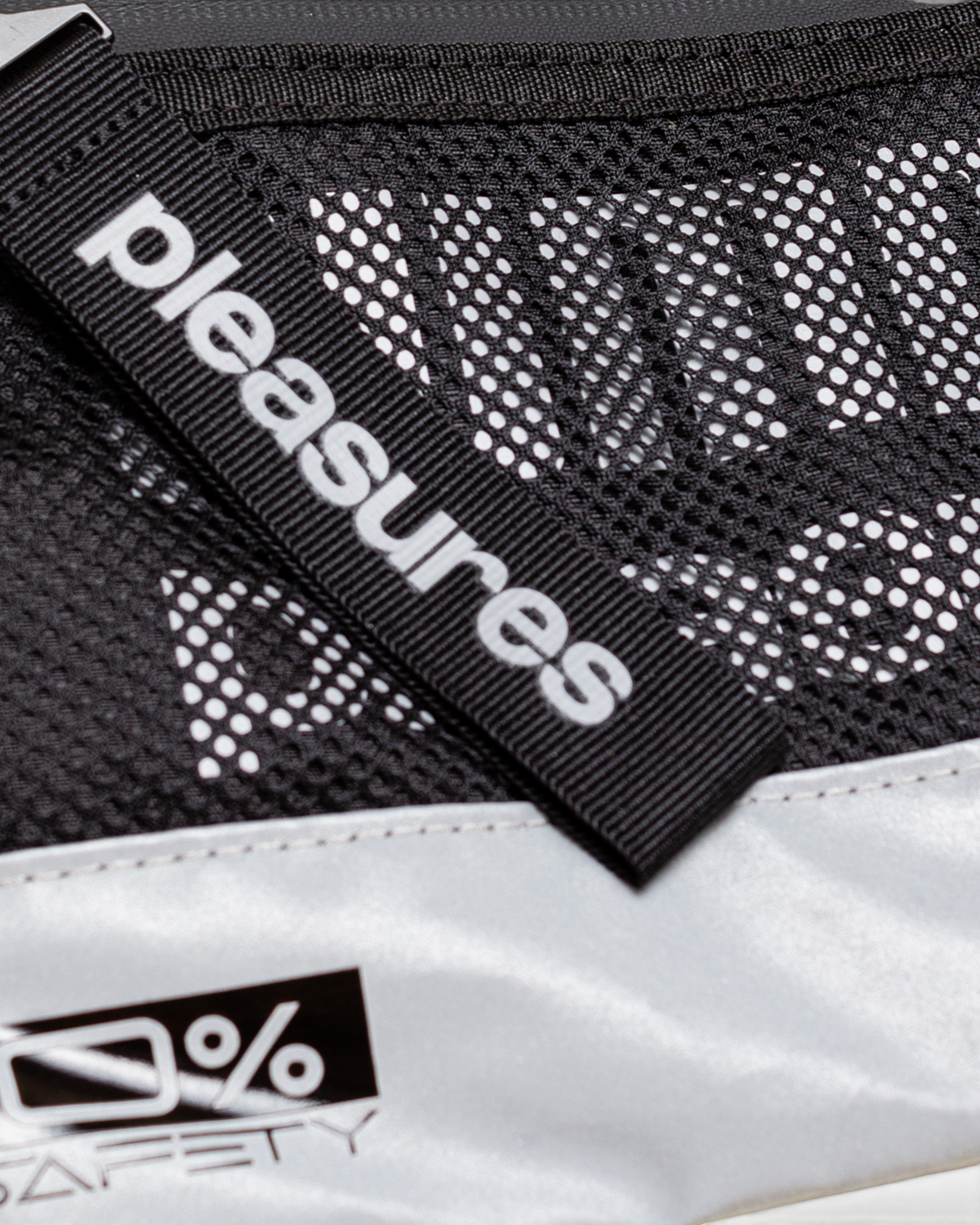 Close up of the Puma x PLEASURES Body Bag in black, the PLEASURES webbing tab lying across the open mesh front panel above the reflective silver base