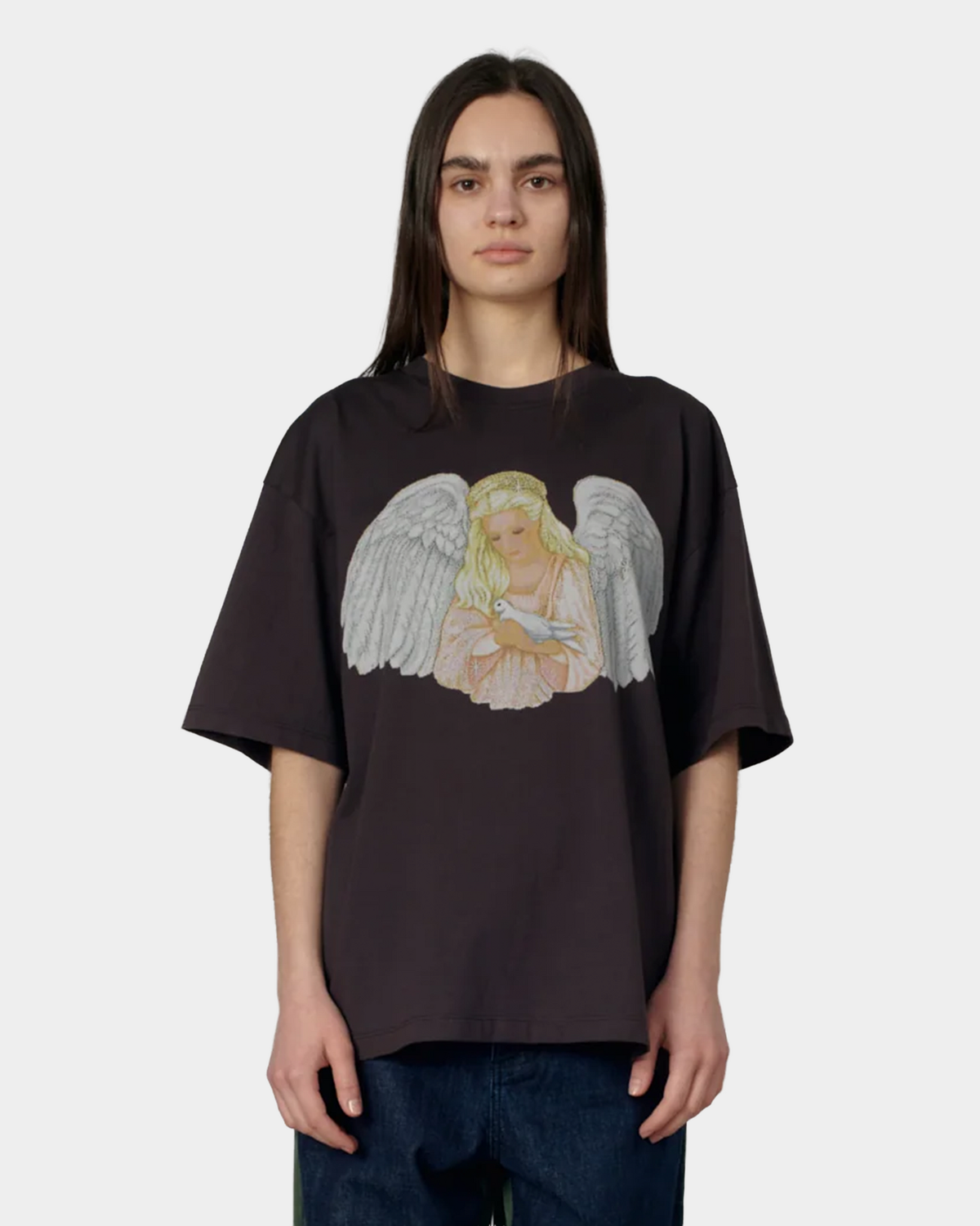 A model with long dark hair wearing the brown Rassvet Blessings T-Shirt loose over dark blue jeans, angel print at the chest