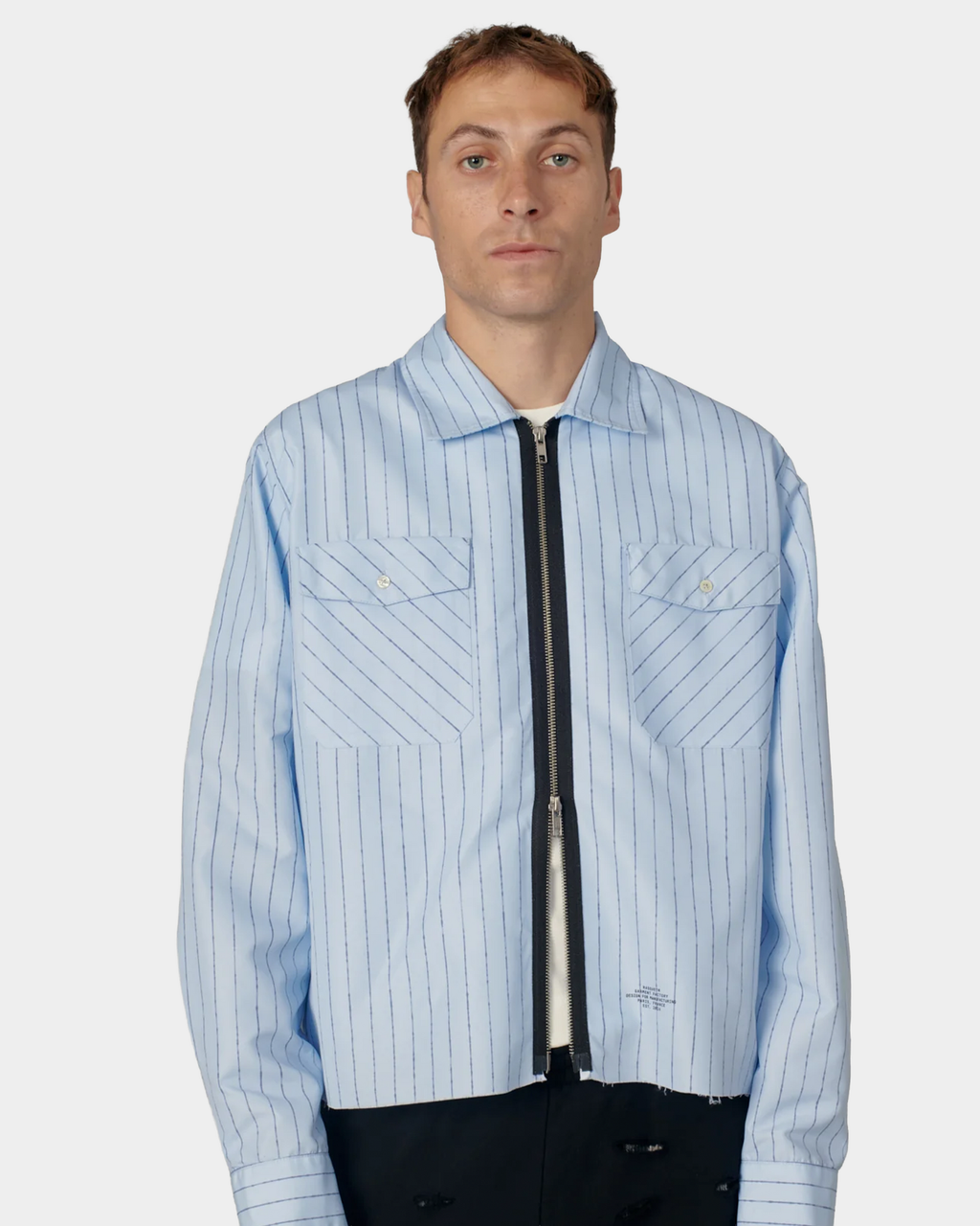 A man wearing the pale blue pinstripe Rassvet Striped Loose Zip Shirt zipped partway, flap chest pockets and buttoned cuffs visible