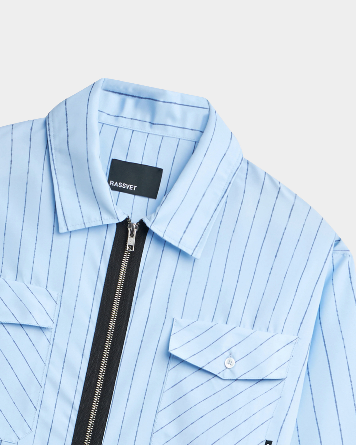 Close-up of the Rassvet Striped Loose Zip Shirt collar and chest, showing the point collar, the exposed black zip tape, a black folded Rassvet label and the pale button on the pocket flap