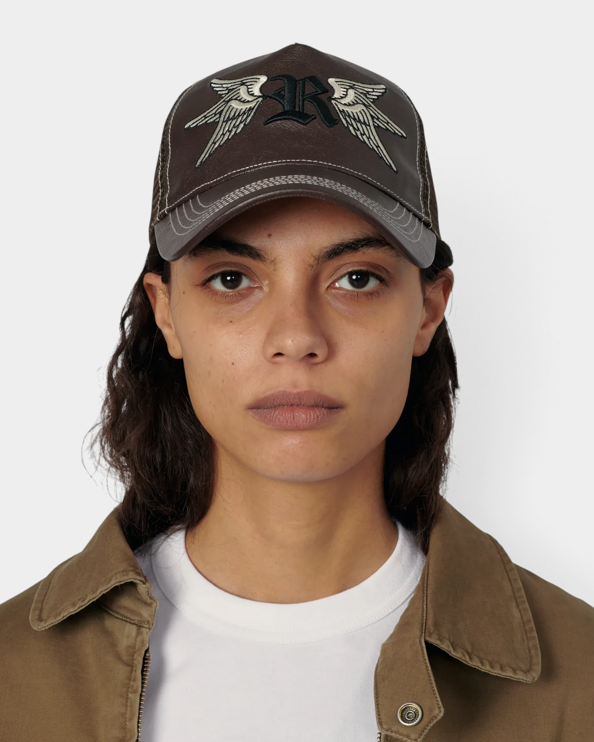 A model with dark hair wearing the brown Rassvet Swallows Trucker Cap over a tan jacket, showing the chocolate brown of the front panel