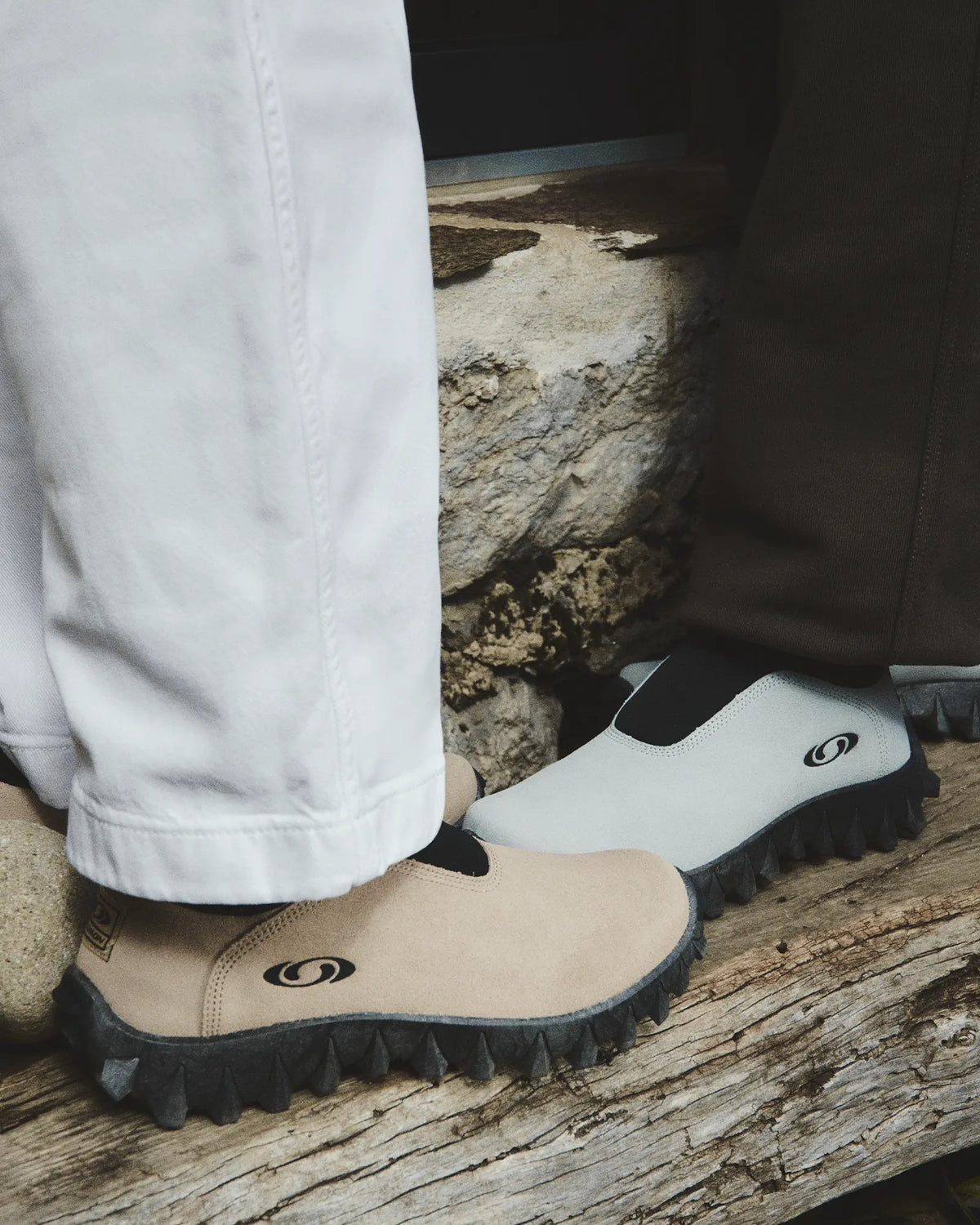 Salomon Snowclog OG Shoes in Safari/Black/Black, close-up of two people's feet on weathered wood, one wearing the shoes in sand, the other in white, above white trousers