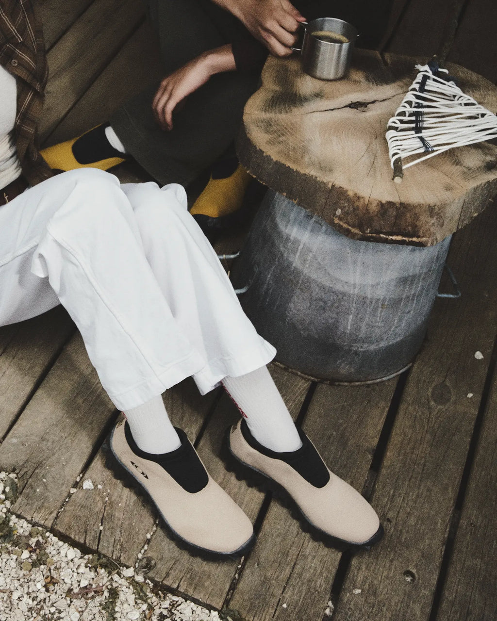 Salomon Snowclog OG Shoes in Safari/Black/Black, person sitting on a wooden deck in white trousers and yellow socks wearing the shoes, beside a metal tub and a tin cup