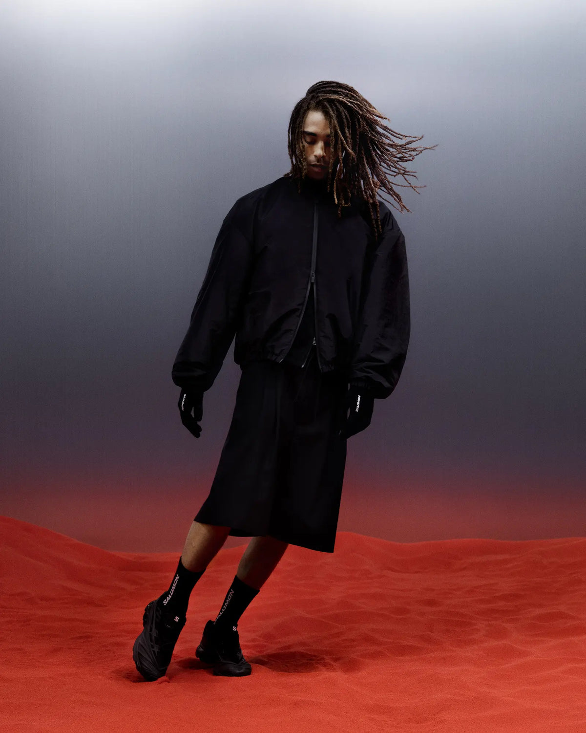 Salomon XT-EVO Shoes in Black/Black/Asphalt, person standing on red ground against a grey backdrop in a black hooded outfit, wearing the shoes