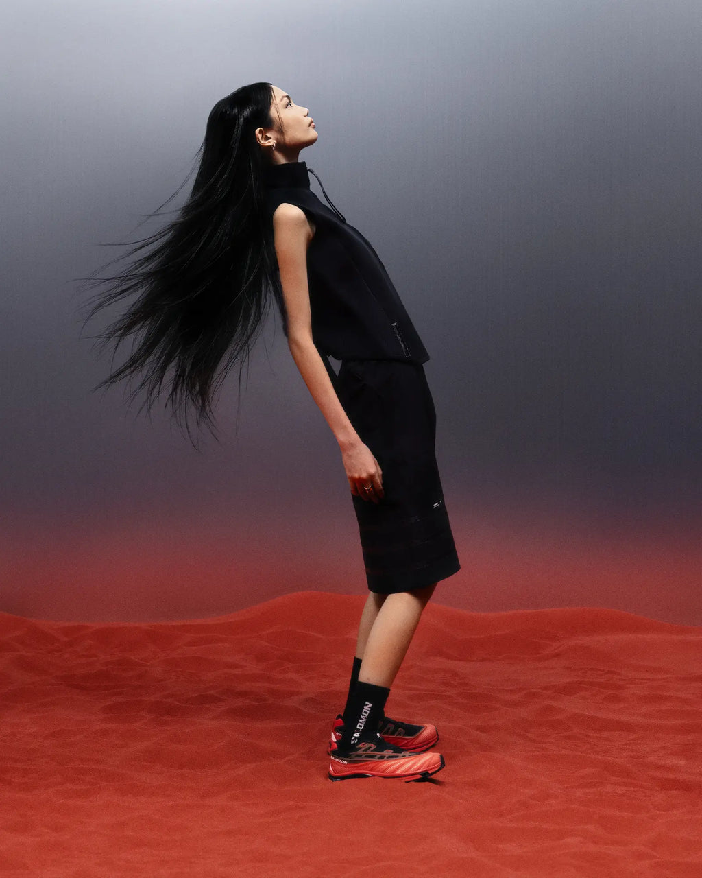 Salomon XT-EVO Shoes in Black/Fiery Red/White, person standing in profile against a red and grey backdrop, long hair loose, dressed in black, wearing the shoes