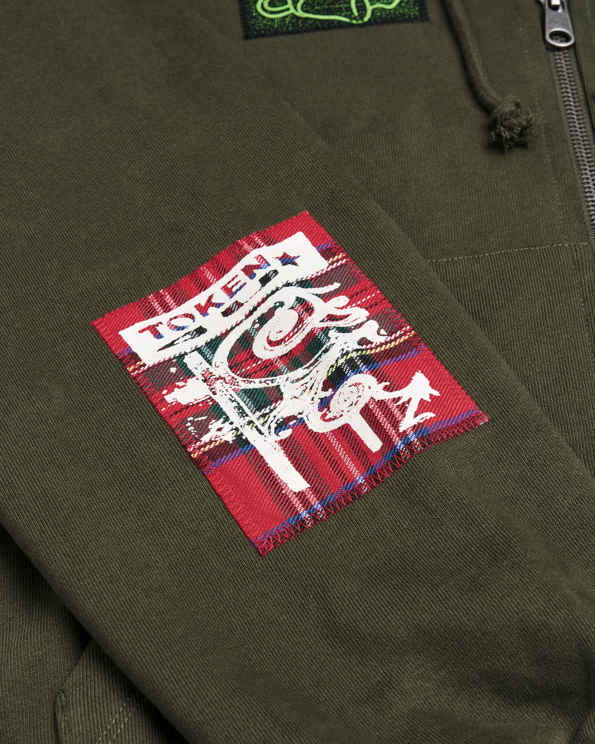 Lockwood Avenue Token Carly zip-up hoodie in olive green with printed and sewn patches, close up of a red patch with white lettering