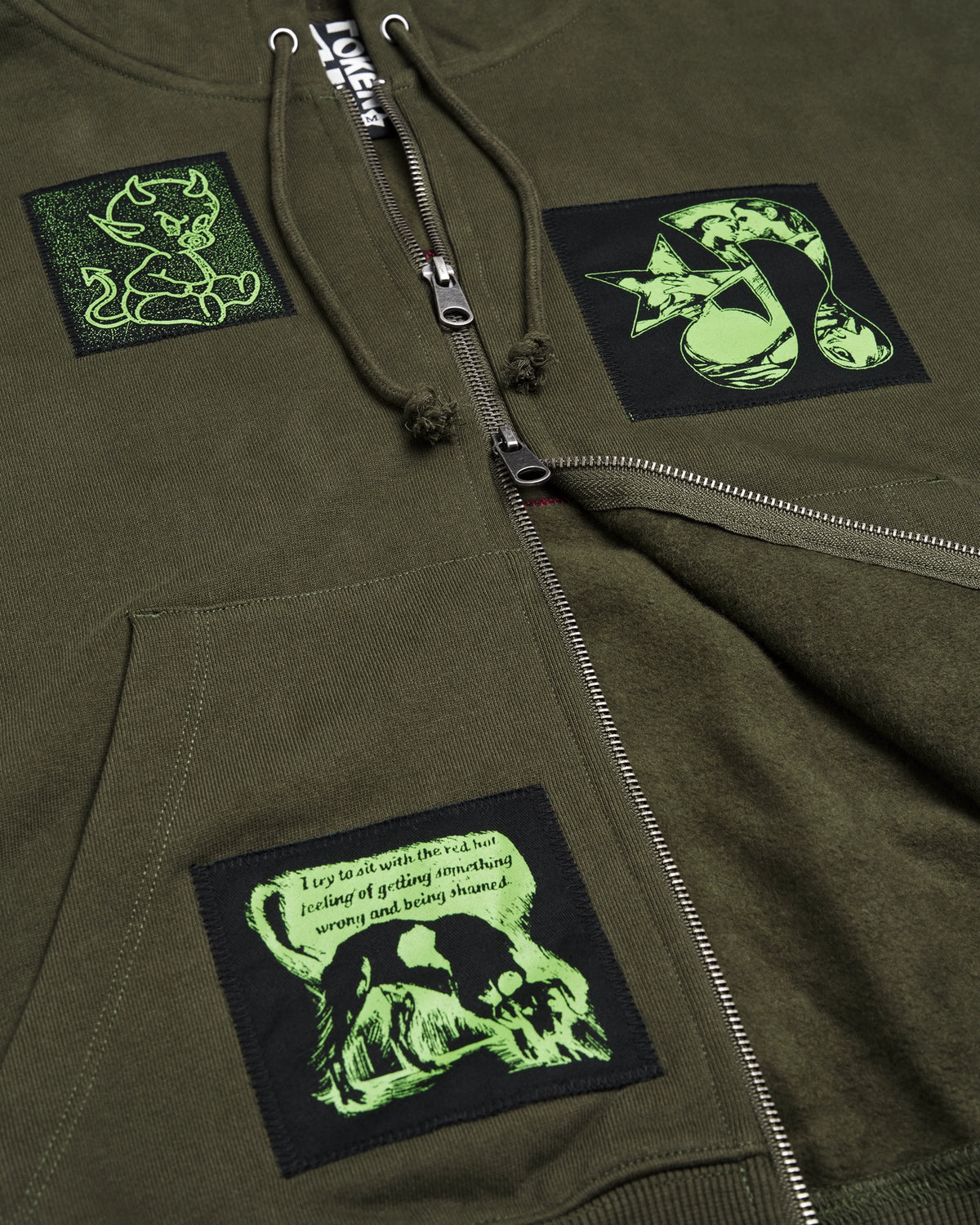 Lockwood Avenue Token Carly zip-up hoodie in olive green with printed and sewn patches, close up of the zip and the patches on the front